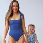 Woman in a blue swimsuit holding hands with a young girl in a floral dress against a plain background
