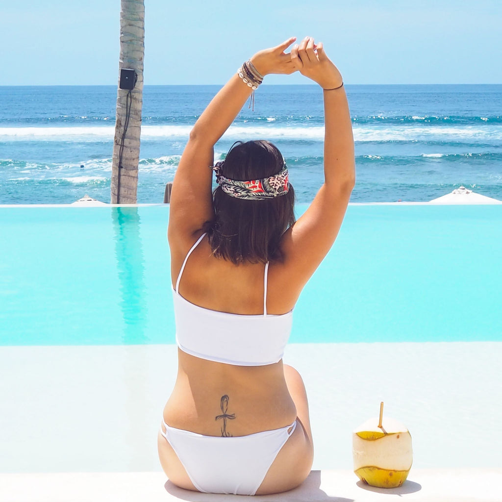 Woman in a white bikini sitting by a pool with ocean view, palm tree, and coconut drink.