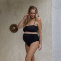 Pregnant Woman wearing a black bikini standing against a neutral wall.