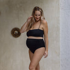 Pregnant Woman wearing a black bikini standing against a neutral wall.