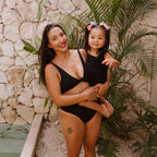 Woman and child in matching black swimsuits standing against a stone wall with greenery.